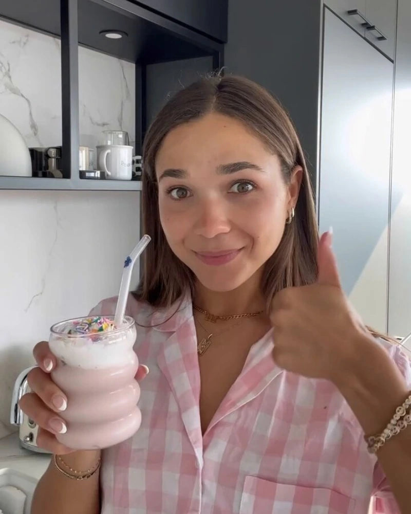 A person in a pink checkered shirt smiles and gives a thumbs up while holding a glass of pink smoothie topped with colorful sprinkles and a straw. They are standing in a modern kitchen.